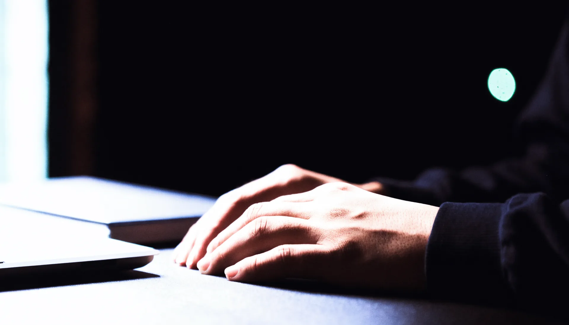 A person's hands at rest on a dark desk beside a closed laptop.